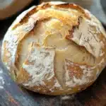 No knead bread with yogurt - the image is a close-up of a freshly baked loaf of bread on a wooden cutting board. the bread is golden brown in color and has a criss-cross pattern on the top. it appears to be freshly baked and is dusted with powdered sugar. the cutting board is placed on a rustic wooden table with a white cloth napkin in the background. the overall mood of the image is warm and inviting.