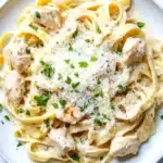 One pot chicken fettuccine - the image is a close-up of a plate of pasta dish. the pasta appears to be fettuccine alfredo, with chunks of chicken and parmesan cheese on top. the dish is garnished with chopped parsley. the plate is white and round, and the pasta is cooked al dente. the background is a white marble countertop.