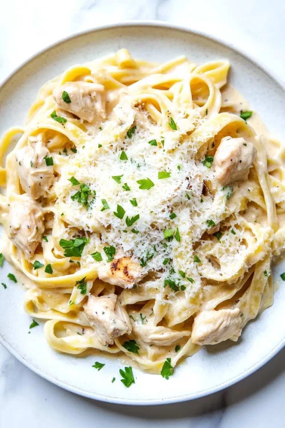 One pot chicken fettuccine - the image is a close-up of a plate of pasta dish. the pasta appears to be fettuccine alfredo, with chunks of chicken and parmesan cheese on top. the dish is garnished with chopped parsley. the plate is white and round, and the pasta is cooked al dente. the background is a white marble countertop.