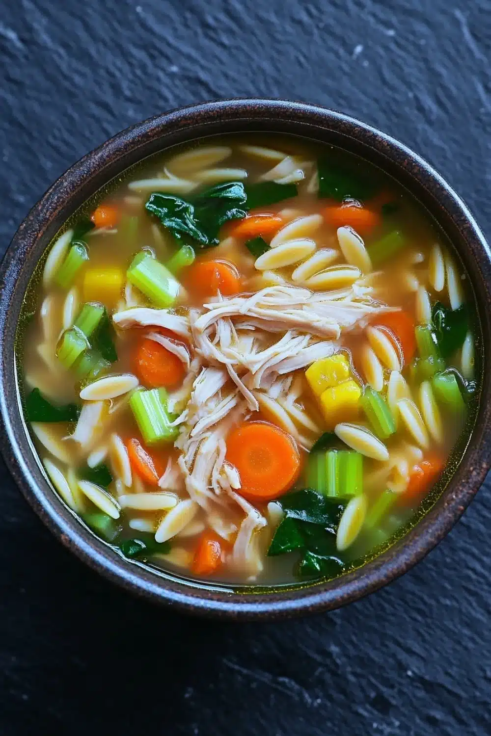 One pot chicken orzo soup - the image is a close-up of a bowl of soup. the soup appears to be a type of noodle soup with chunks of chicken, carrots, celery, and other vegetables mixed in. the bowl is made of dark brown ceramic and is sitting on a dark grey surface. the broth is a light yellow color and looks thick and creamy. there are also some green leaves scattered throughout the soup, adding a pop of color to the dish.