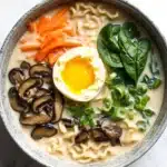 One pot creamy ramen - the image is a close-up of a bowl of ramen soup. the bowl is made of ceramic and is sitting on a white marble countertop. the ramen is a light beige color and appears to be thick and creamy. on top of the ramen, there is a hard-boiled egg with a runny yolk. next to the egg, there are sliced carrots, sliced mushrooms, and fresh green onions. the vegetables are arranged in a way that they are overlapping each other. the overall appearance of the dish is fresh and appetizing.