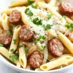One pot garlic sausage pasta - the image is a close-up of a bowl of pasta dish. the pasta appears to be penne, which is a type of penne pasta, and it is cooked al dente. the dish is garnished with chopped parsley and grated parmesan cheese. there are also small chunks of sausage scattered throughout the dish, which are golden brown and appear to be seasoned with herbs and spices. the bowl is white and is sitting on a white marble countertop.