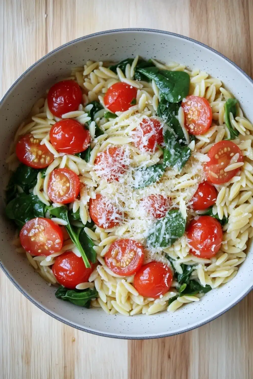 One pot pasta orzo - the image is a close-up of a bowl of pasta salad. the bowl is white with a blue rim and is sitting on a wooden table. the pasta is fusilli, which is a type of spiral-shaped pasta, and it is mixed with cherry tomatoes, spinach, and parmesan cheese. the tomatoes are bright red and appear to be ripe and juicy. the spinach is a vibrant green color and is scattered throughout the dish. the cheese is grated and is sprinkled on top of the pasta. the overall appearance of the dish is fresh and appetizing.