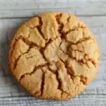 Peanut butter cookie no butter - the image is a close-up of a round cookie on a white textured surface. the cookie appears to be freshly baked and has a golden-brown color. it has a crumbly texture on the surface, with small pieces of peanut butter scattered around it. the background is blurred, making the cookie the focal point of the image.