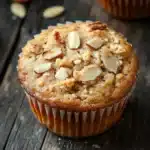 Protein muffin almond flour - the image shows a close-up of a freshly baked muffin on a wooden table. the muffin is golden brown in color and has a crumbly texture. it is topped with sliced almonds, giving it a crunchy appearance. there are a few scattered almonds around the muffin, adding a pop of color to the image. the background is blurred, but it appears to be a rustic wooden surface.