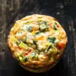 Protein veggie muffin - the image is a close-up of a round muffin on a dark wooden surface. the muffin appears to be freshly baked and has a golden brown crust. it is filled with a mixture of vegetables, including zucchini, red bell peppers, and green onions. the vegetables are scattered throughout the mixture, with some of them overlapping each other. the top of the muffin is covered in melted cheese, which is a light yellow color. the background is blurred, making the muffins the focal point of the image.