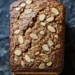 Pumpkin bread almond flour - the image is a close-up of a rectangular loaf of bread. the bread appears to be freshly baked and has a golden brown color. it is topped with sliced almonds, giving it a crunchy texture. the loaf is resting on a dark, textured surface, possibly a table or countertop. the overall mood of the image is warm and inviting.