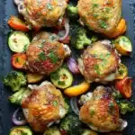 Sheet pan boneless skinless chicken thighs and vegetables - the image shows a tray of grilled chicken and vegetables. the chicken is golden brown and appears to be seasoned with herbs and spices. the vegetables are arranged in a circular pattern on the tray, with some overlapping each other. there are also small pieces of broccoli, zucchini, and red onions scattered throughout the tray. the tray is black and there are a few sprigs of parsley on the side. the overall presentation of the dish is colorful and appetizing.