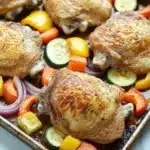 Sheet pan chicken and veggies - the image shows a baking tray with four whole chicken breasts on it. the chicken breasts are golden brown and appear to be seasoned with herbs and spices. the tray is filled with sliced vegetables, including zucchini, red onions, and yellow bell peppers. the vegetables are arranged in a circular pattern on the tray, with some overlapping each other. the background is a white marble countertop.