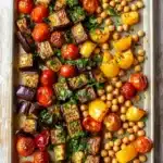 Sheet pan dinner eggplant - the image shows a rectangular baking tray filled with roasted vegetables. the vegetables are arranged in a single layer on the tray, with some of them overlapping each other. the tray is lined with parchment paper and is placed on a wooden table. the vegetables appear to be grilled eggplant, cherry tomatoes, and yellow bell peppers. the eggplant is cut into small cubes and is garnished with chopped parsley. the cherry tomatoes are bright red and appear to have been roasted to perfection, while the bell peppers are yellow and have a slightly charred texture. the chickpeas are scattered throughout the tray in a pile, adding a pop of color to the dish. the overall appearance of the dish is colorful and appetizing.