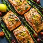 Sheet pan lemon brown butter salmon - the image shows three pieces of salmon fillets on a black slate plate. the salmon is cooked to a golden brown color and is garnished with herbs and lemon zest. the plate is also decorated with cherry tomatoes and asparagus spears. there is a lemon wedge on the side of the plate, adding a pop of color to the dish. the overall presentation is elegant and appetizing.