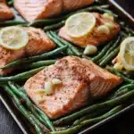 Sheet pan salmon green beans - the image shows a baking tray with four pieces of salmon and green beans. the salmon is cooked to a golden brown color and appears to be seasoned with herbs and spices. the green beans are long and slender, and they are arranged in a single layer on the tray. there are also slices of lemon on top of the salmon, adding a pop of color to the dish. the tray is placed on a wooden table, and there are a few plates visible in the background.