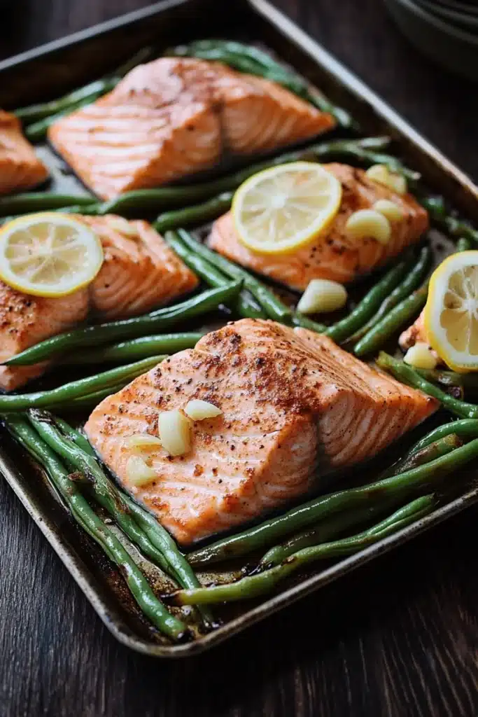 Sheet pan salmon green beans - the image shows a baking tray with four pieces of salmon and green beans. the salmon is cooked to a golden brown color and appears to be seasoned with herbs and spices. the green beans are long and slender, and they are arranged in a single layer on the tray. there are also slices of lemon on top of the salmon, adding a pop of color to the dish. the tray is placed on a wooden table, and there are a few plates visible in the background.