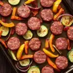 Sheet pan smoked sausage and veggies - the image shows a baking tray filled with various types of sausage and vegetables. the tray is lined with parchment paper and is placed on a wooden table. the sausage is arranged in a circular pattern on the tray, with some overlapping each other. there are also slices of zucchini, red onions, and yellow bell peppers scattered throughout the tray. the vegetables appear to be seasoned with herbs and spices, giving them a golden brown color. the background is blurred, but it appears to be a kitchen countertop.