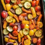 Sheet pan vegetables air fryer - the image shows a baking tray filled with roasted vegetables. the vegetables are arranged in a circular pattern on the tray, with some overlapping each other. the colors of the vegetables include yellow, orange, red, and green bell peppers, as well as sliced red onions. the tray is lined with parchment paper, and there is a green cloth napkin on the side. the background is a wooden table.