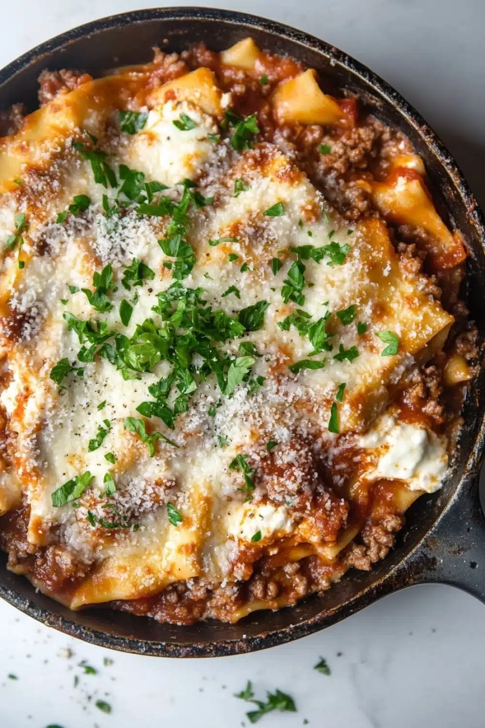 Skillet beef lasagna - the image shows a close-up of a dish in a black cast iron skillet. the dish appears to be a lasagna or lasagna, with layers of layers of meat and cheese on top. the meat is a rich, dark brown color, and the cheese is melted and bubbly. there are also a few sprigs of fresh parsley scattered on top, adding a pop of green color to the dish. the skillet is sitting on a white marble countertop.