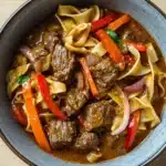 Slow cooker beef noodle curry - the image is a close-up of a bowl of beef noodle soup. the bowl is made of ceramic and has a light blue rim. the soup is a rich brown color and appears to be thick and creamy. it is filled with chunks of beef, red and orange bell peppers, onions, and noodles. the noodles are thin and appear to be cooked until they are soft and fluffy. there are also small pieces of green herbs scattered throughout the soup, adding a pop of color to the dish. the dish is garnished with a sprig of parsley.