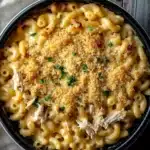 Slow cooker chicken mac and cheese - the image shows a black bowl filled with a creamy macaroni and cheese dish. the dish is topped with a layer of breadcrumbs and sprinkled with chopped parsley. the bowl is sitting on a wooden table with a white cloth napkin on the side. the macaronis are long and thin, and the cheese is a light yellow color. there are also chunks of chicken visible in the dish, which appears to be cooked until it is golden brown. the overall appearance of the dish is savory and appetizing.