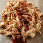 Slow cooker chicken pulled - the image shows a pile of shredded chicken on a wooden cutting board. the chicken appears to be cooked and has a golden brown color. it is covered in a thick, dark sauce that is dripping down the sides of the chicken. the sauce is thick and thick, and it looks like it would be a type of barbecue sauce. the cutting board has a light-colored wood grain pattern, and the background is blurred, making the chicken the focal point of the image.
