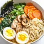 Slow cooker chicken ramen - the image is a close-up of a bowl of ramen. the bowl is white and is sitting on a white marble countertop. the ramen is made up of noodles, sliced carrots, sliced mushrooms, sliced chicken, sliced green onions, and a hard-boiled egg. the noodles are light brown and appear to be thick and fluffy. there are also some sesame seeds sprinkled on top of the noodles. the dish is garnished with a sprinkle of sesame seed.