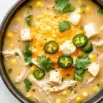 Slow cooker jalapeno popper chicken soup - the image is a close-up of a bowl of soup. the soup appears to be a creamy yellow color with chunks of chicken, corn, and jalapenos scattered throughout. the bowl is made of dark brown ceramic and is sitting on a white marble countertop. there are also a few sprigs of cilantro scattered around the bowl. the overall appearance of the soup is fresh and appetizing.