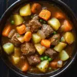 Slow cooker poor mans beef stew - the image is a close-up of a bowl of beef stew. the stew is made with chunks of beef, potatoes, carrots, and peas. the beef is cooked in a rich, dark brown broth and is garnished with chopped parsley. the potatoes are yellow and appear to be diced, while the carrots are orange and the peas are green. the bowl is black and is sitting on a dark wooden table. there are a few sprigs of parsley scattered around the bowl.