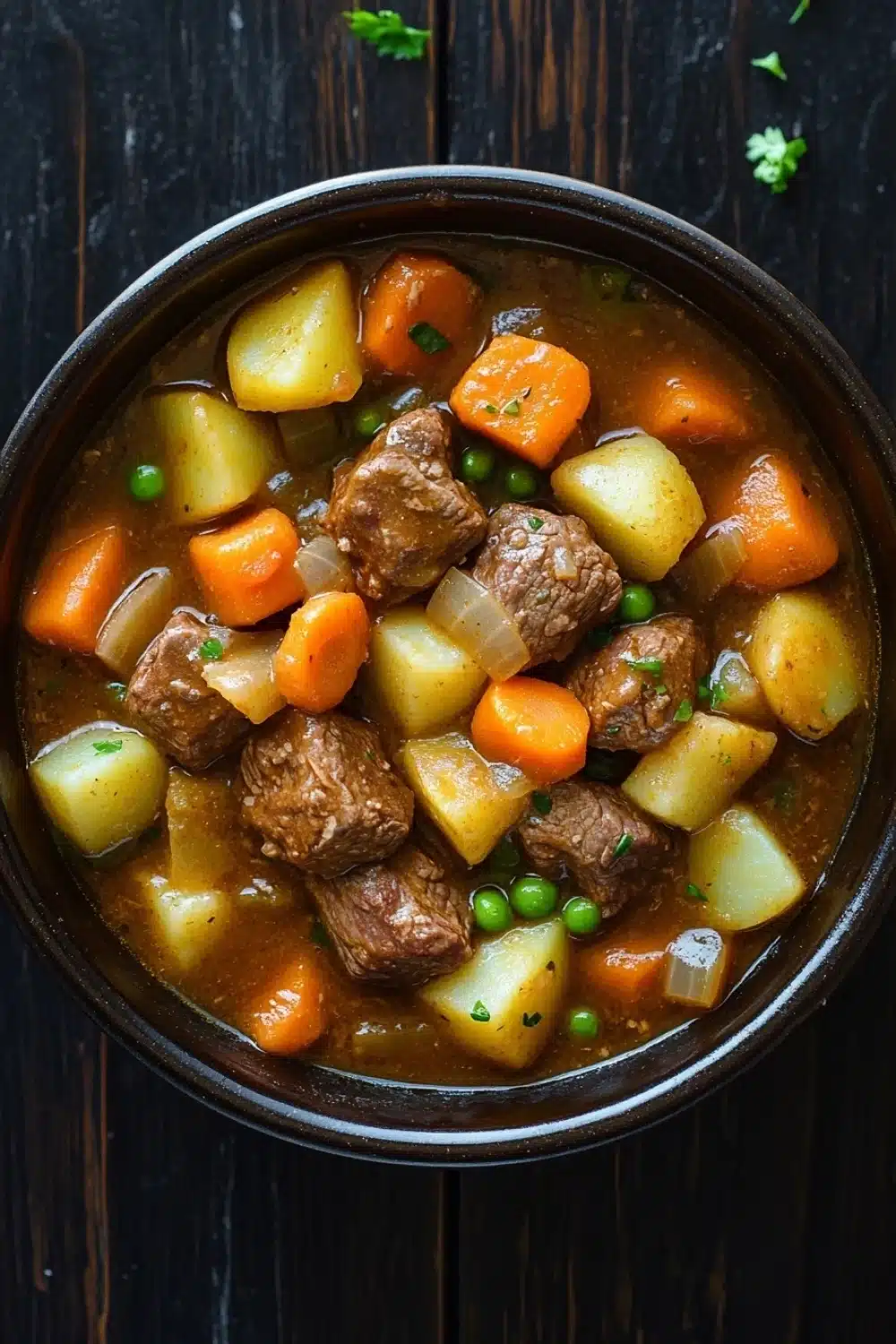 Slow cooker poor mans beef stew - the image is a close-up of a bowl of beef stew. the stew is made with chunks of beef, potatoes, carrots, and peas. the beef is cooked in a rich, dark brown broth and is garnished with chopped parsley. the potatoes are yellow and appear to be diced, while the carrots are orange and the peas are green. the bowl is black and is sitting on a dark wooden table. there are a few sprigs of parsley scattered around the bowl.