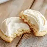Sugar cookie and frosting - the image shows two cookies on a wooden table. the cookies are round and golden brown in color, with a light dusting of white frosting on top. one of the cookies has a bite taken out of it, revealing the soft, fluffy interior. the background is blurred, but it appears to be a kitchen countertop with a white cloth napkin on the side. the overall mood of the image is warm and inviting.