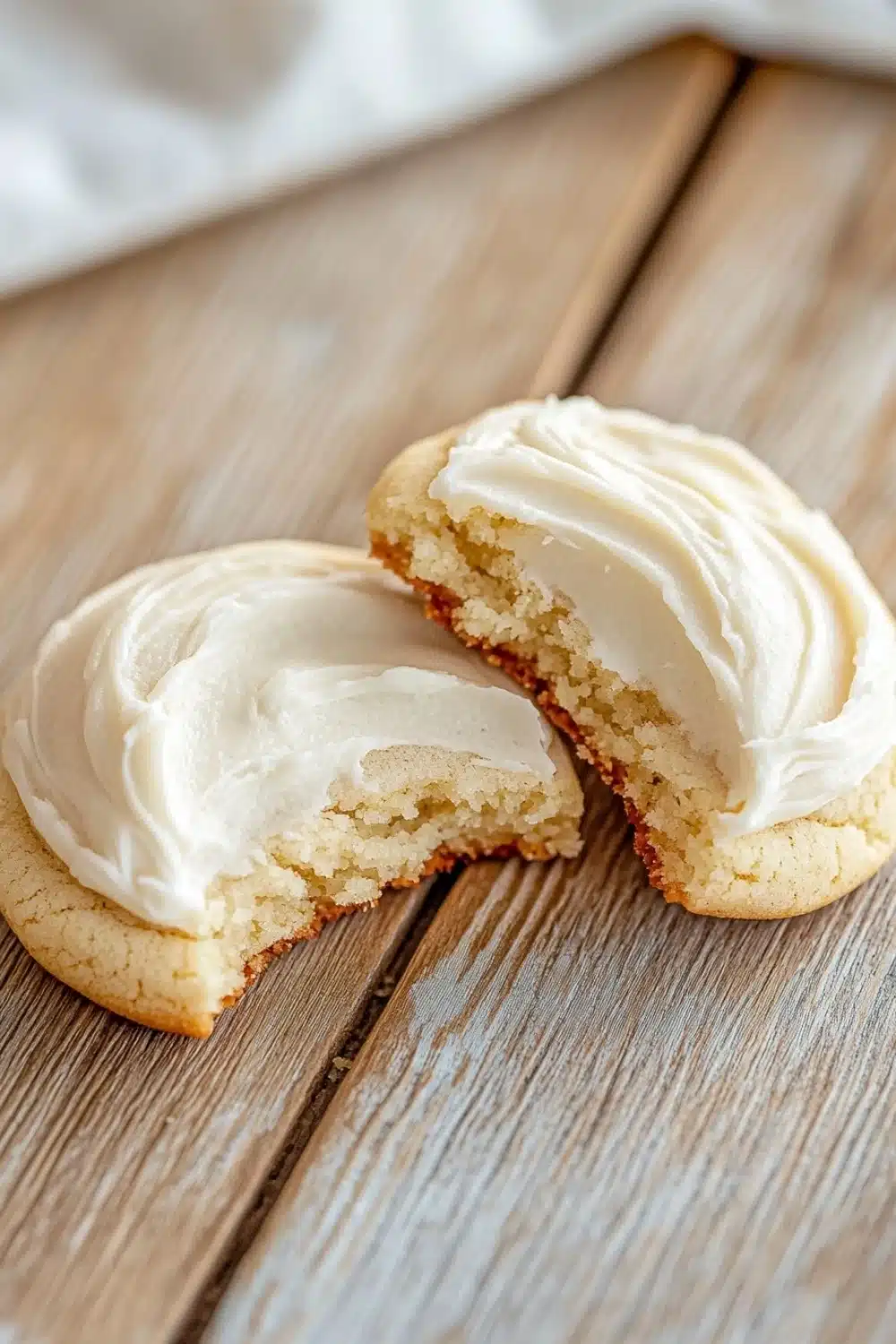 Sugar cookie and frosting - the image shows two cookies on a wooden table. the cookies are round and golden brown in color, with a light dusting of white frosting on top. one of the cookies has a bite taken out of it, revealing the soft, fluffy interior. the background is blurred, but it appears to be a kitchen countertop with a white cloth napkin on the side. the overall mood of the image is warm and inviting.