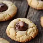 Sugar cookie kisses - the image shows a group of freshly baked cookies on a wooden surface. the cookies are round and golden brown in color, with a crumbly texture. they are arranged in a scattered manner, with some overlapping each other. in the center of the image, there is a single chocolate chip cookie with a bite taken out of it. the chocolate chip is covered in a thick layer of dark chocolate. the background is blurred, but it appears to be a rustic wooden table.