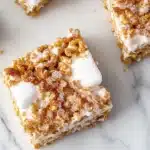 Sugar cookie rice crispy - the image shows three square-shaped dessert bars on a white marble surface. the bars are made with a layer of white chocolate and topped with a generous amount of crumbled granola. the granola is a light brown color and appears to be drizzled over the top of the bars. there are also a few small pieces of marshmallows scattered around the bars, adding a pop of color to the image.