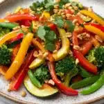 Thai vegetable air fryer - the image is a close-up of a colorful salad in a round plate. the salad is made up of various types of vegetables, including bell peppers, zucchini, broccoli, and pine nuts. the bell peppers are bright red and yellow, while the broccoli is green and the pine nuts are scattered throughout the salad. there are also some sprigs of cilantro on top of the vegetables. the plate is sitting on a white marble countertop.