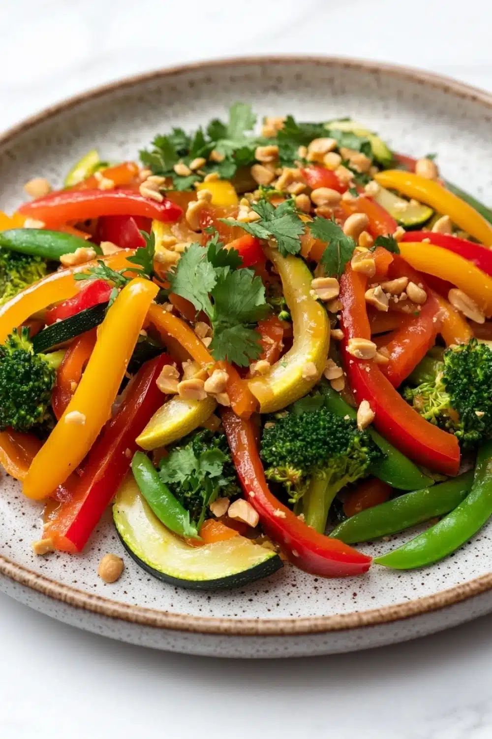 Thai vegetable air fryer - the image is a close-up of a colorful salad in a round plate. the salad is made up of various types of vegetables, including bell peppers, zucchini, broccoli, and pine nuts. the bell peppers are bright red and yellow, while the broccoli is green and the pine nuts are scattered throughout the salad. there are also some sprigs of cilantro on top of the vegetables. the plate is sitting on a white marble countertop.