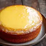 Yellow lemon cake design - the image is a close-up of a round cake on a white plate. the cake appears to be a cheesecake with a golden brown crust and a creamy yellow filling. the filling is spread evenly across the top of the cake, creating a smooth and creamy texture. the plate is placed on a wooden table with a rustic background. the overall mood of the image is warm and inviting.